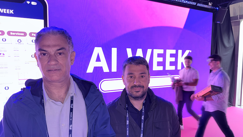 Two NETSQUARE representatives wearing AI WEEK lanyards stand in front of a large illuminated AI WEEK screen at the event.