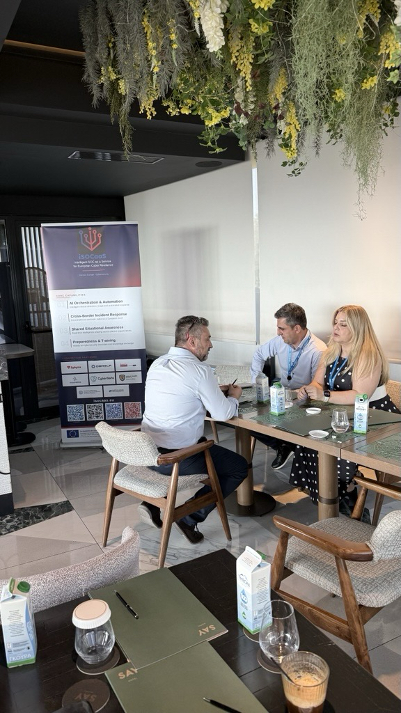 Participants in discussion at a table next to the iSOCaaS roll-up banner presenting the project’s core capabilities, at Hotel SAY in Athens.